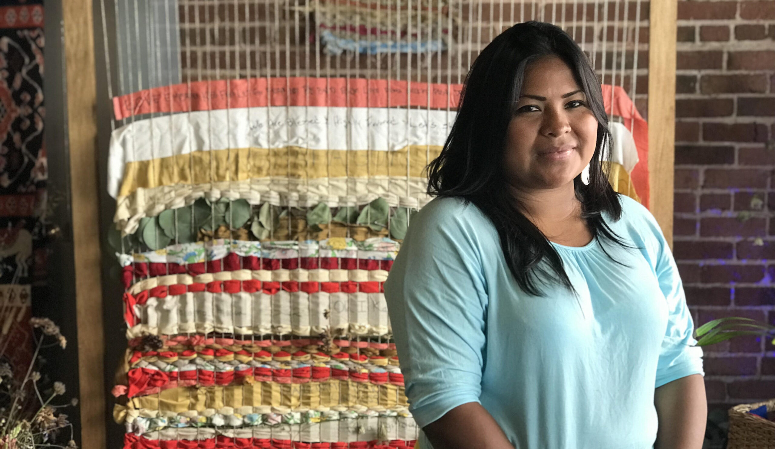 Foto colorida de Sara Omi. Ela é uma indígena Emberá com cabelos lisos pretos. Ela usa uma camiseta azul clara e ao fundo aparece uma tapeçaria colorida