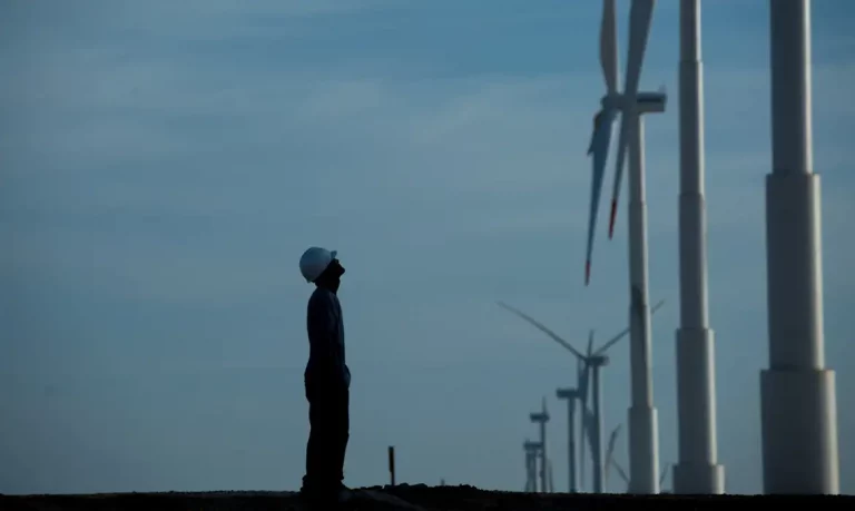 Foto colorida do perfil de um homem de pé diante de turbina de energia eólica. Ao fundo, céu azul