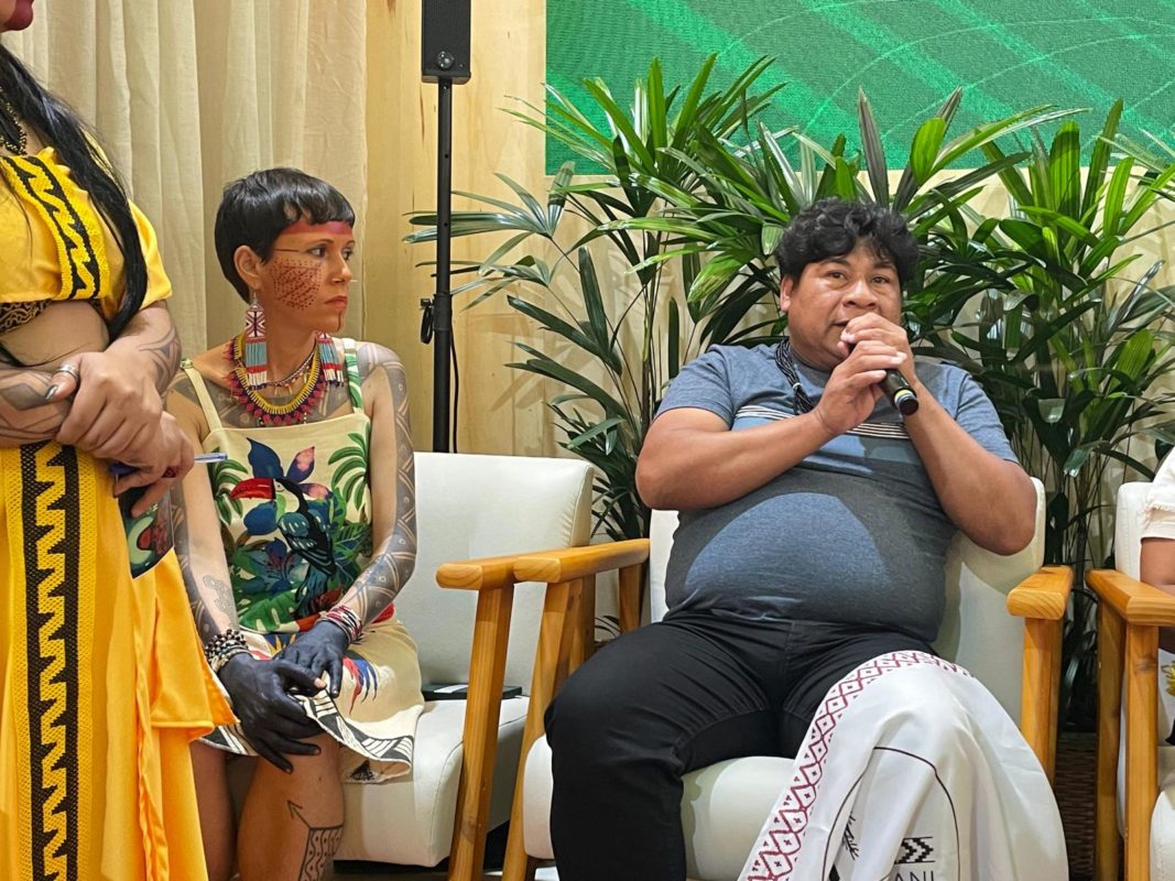 Foto colorida de Leonardo Werá Tupã e Clara Opoxina. Eles estão sentados e Leonardo segura um microfone. Estão em painel sobre danos espirituais na COP30