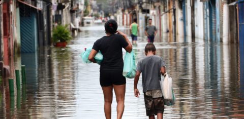 Foto colorida de pessoas andando em meio a rua alagada na zona leste de São Paulo após enchente. Situação revela relação entre clima e saúde