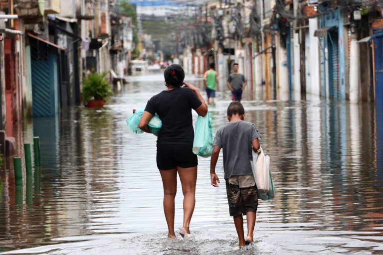 Foto colorida de pessoas andando em meio a rua alagada na zona leste de São Paulo após enchente. Situação revela relação entre clima e saúde