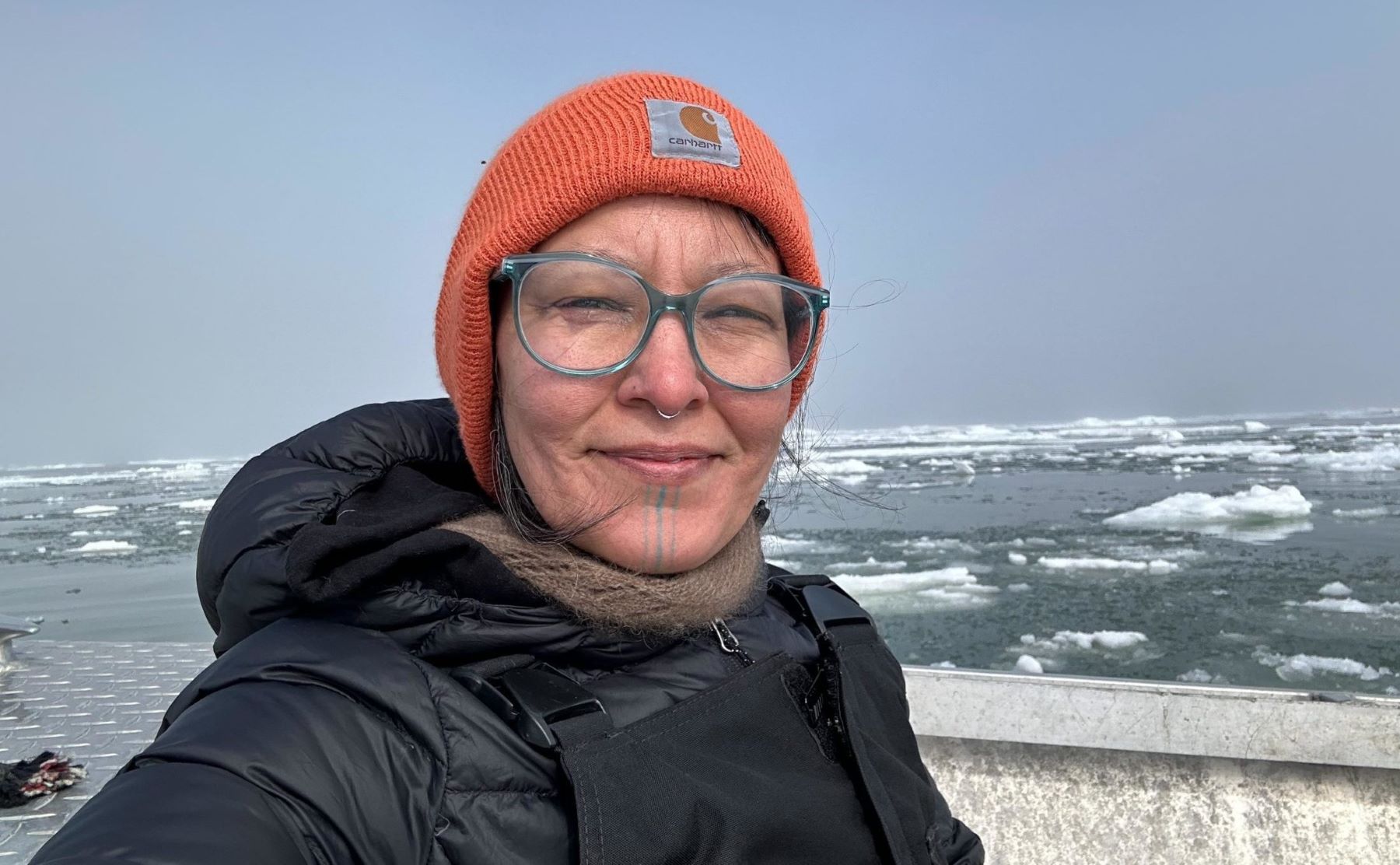 Foto colorida de Aakaluk em Unalakleet, pequena cidade costeira do distrito de Nome, no Alasca, Estados Unidos. Na imagem, ela aparece de casaco preto e toca laranja. Ao fundo, geleira derretendo