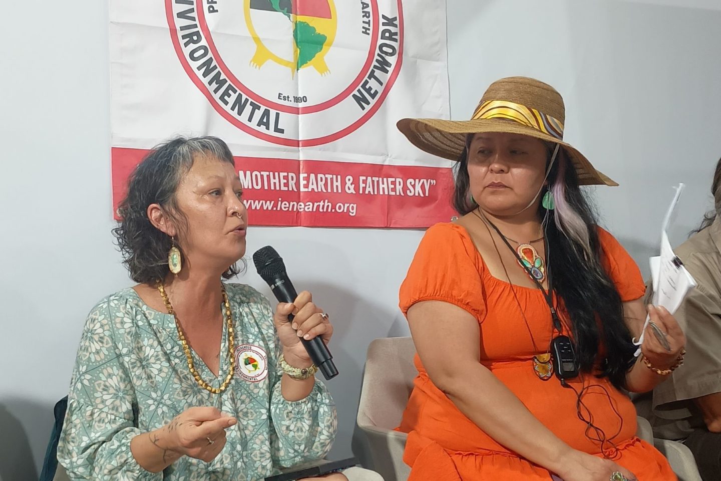 Foto colorida de Aakaluk, nome nativo de Adrienne La-Ree Blatchford. Ela é uma mulher indígena de cabelos curtos pretos e meio grisalhos. Ela está com um vestido verde e segura um microfone