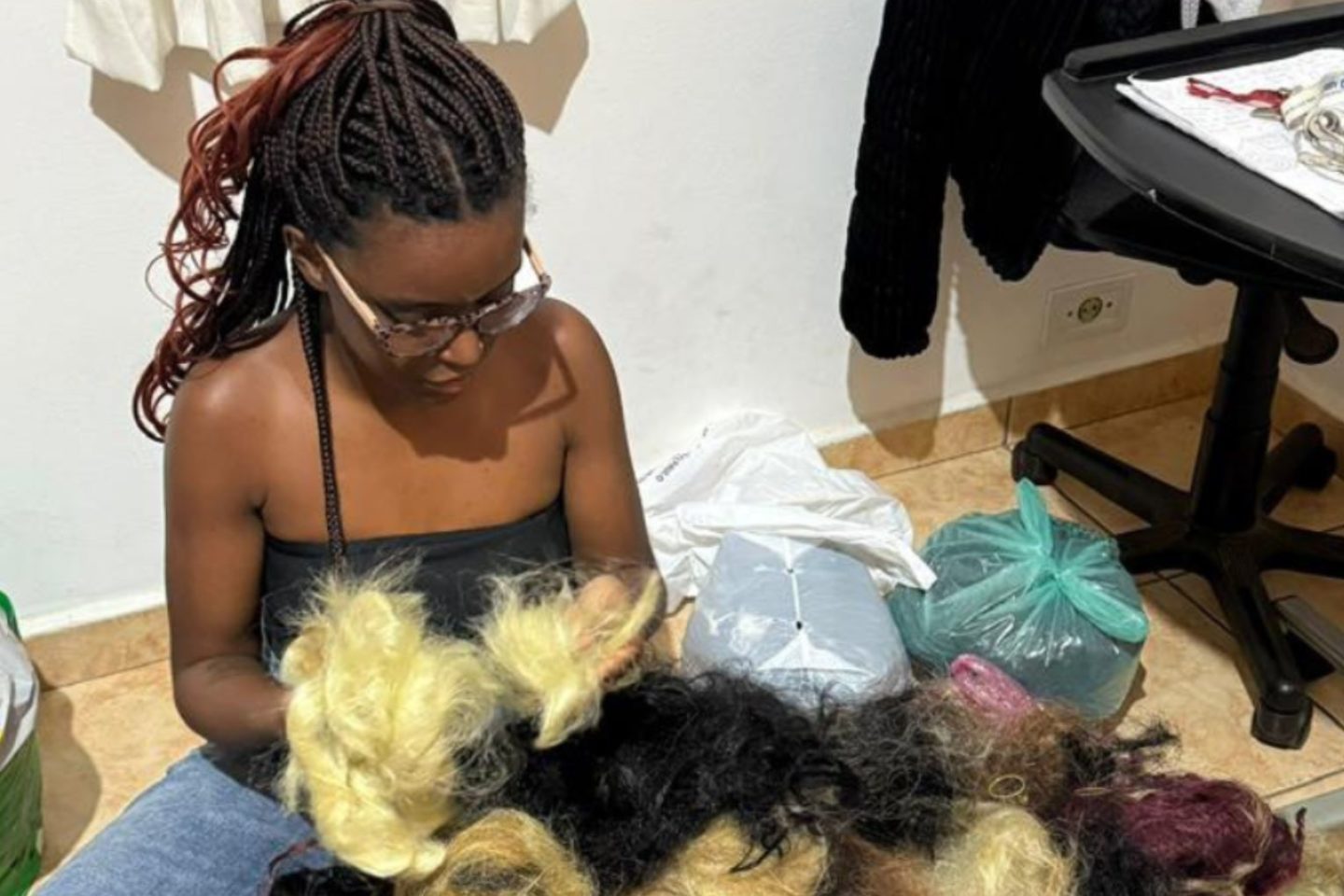 Foto colorida de Vitória Emanoely Campos. Ela é uma mulher preta com tranças afro pretas e está sentada diante de vários pedaços de cabelo sintético. Trancista criou o Recicla Braids