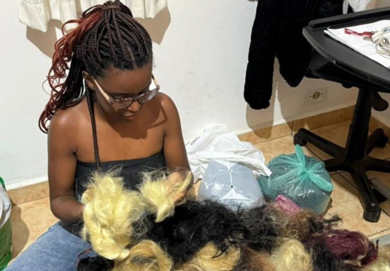 Foto colorida de Vitória Emanoely Campos. Ela é uma mulher preta com tranças afro pretas e está sentada diante de vários pedaços de cabelo sintético. Trancista criou o Recicla Braids