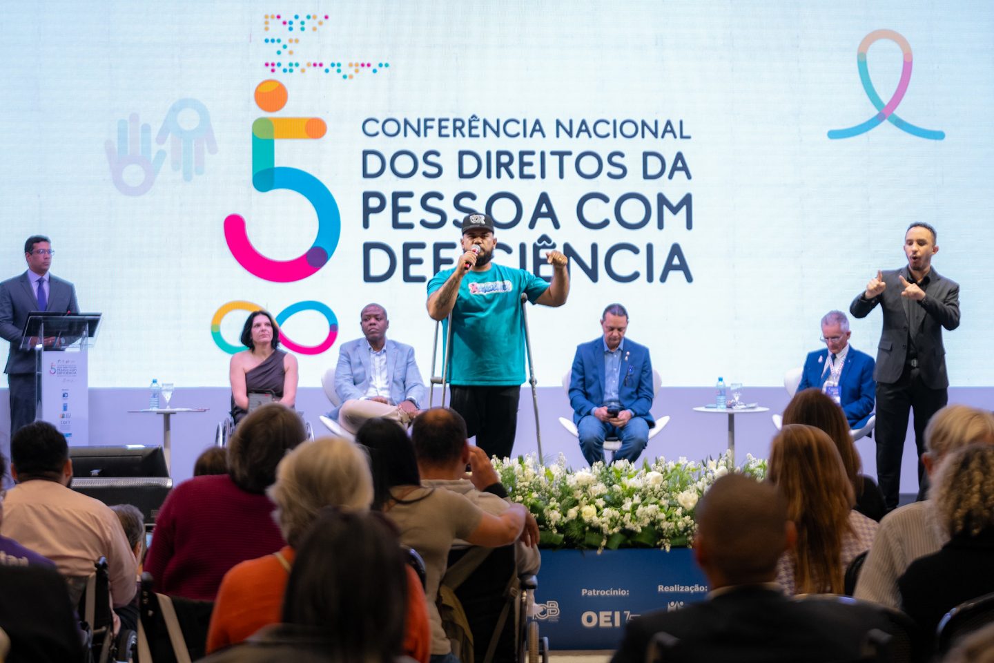 Foto colorida de manifestação de uma pessoa com deficiência durante a 5° Conferência Nacional dos Direitos da Pessoa com Deficiência. Na imagem, o homem negro usa muletas e segura um microfone, enquanto fala e gesticula com o braço esquerdo levantado, enquanto olha para a plateia que está à sua frente. Ao fundo, pessoas sentadas no palco.
