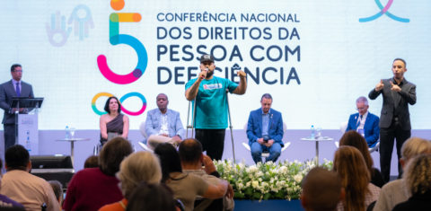 Foto colorida de manifestação de uma pessoa com deficiência durante a 5° Conferência Nacional dos Direitos da Pessoa com Deficiência. Na imagem, o homem negro usa muletas e segura um microfone, enquanto fala e gesticula com o braço esquerdo levantado, enquanto olha para a plateia que está à sua frente. Ao fundo, pessoas sentadas no palco.