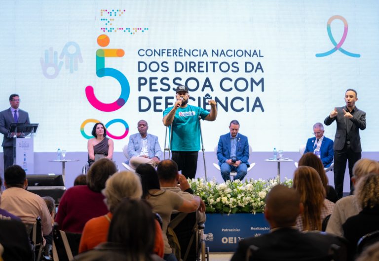 Foto colorida de manifestação de uma pessoa com deficiência durante a 5° Conferência Nacional dos Direitos da Pessoa com Deficiência. Na imagem, o homem negro usa muletas e segura um microfone, enquanto fala e gesticula com o braço esquerdo levantado, enquanto olha para a plateia que está à sua frente. Ao fundo, pessoas sentadas no palco.