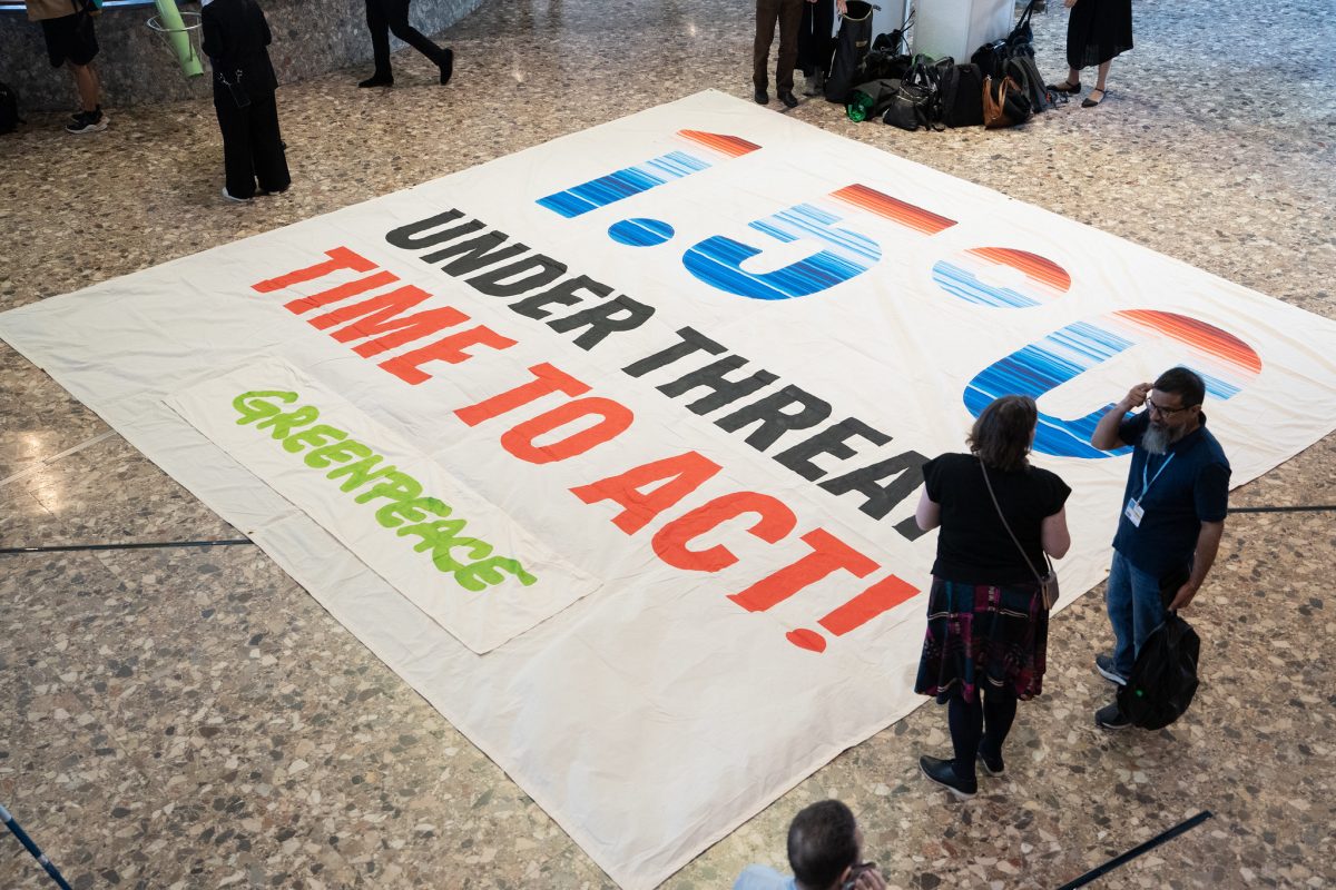 Foto colorida de cartaz colocado no chão na Conferência de Bonn. No cartaz aparece a temperatura 1,5°C e a frase em inglês: Under Threat Time To Act!. Abaixo aparece o logo do Greenpeace e a cor do banner é branca com letras coloridas