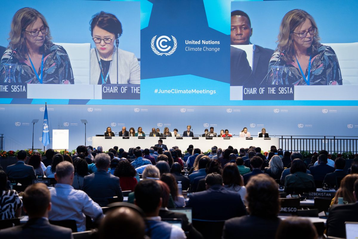Foto colorida das discussões da Conferência de Bonn. Na imagem, aparece a plateia e uma mesa com autoridades. Ao fundo, telão com algumas pessoas.