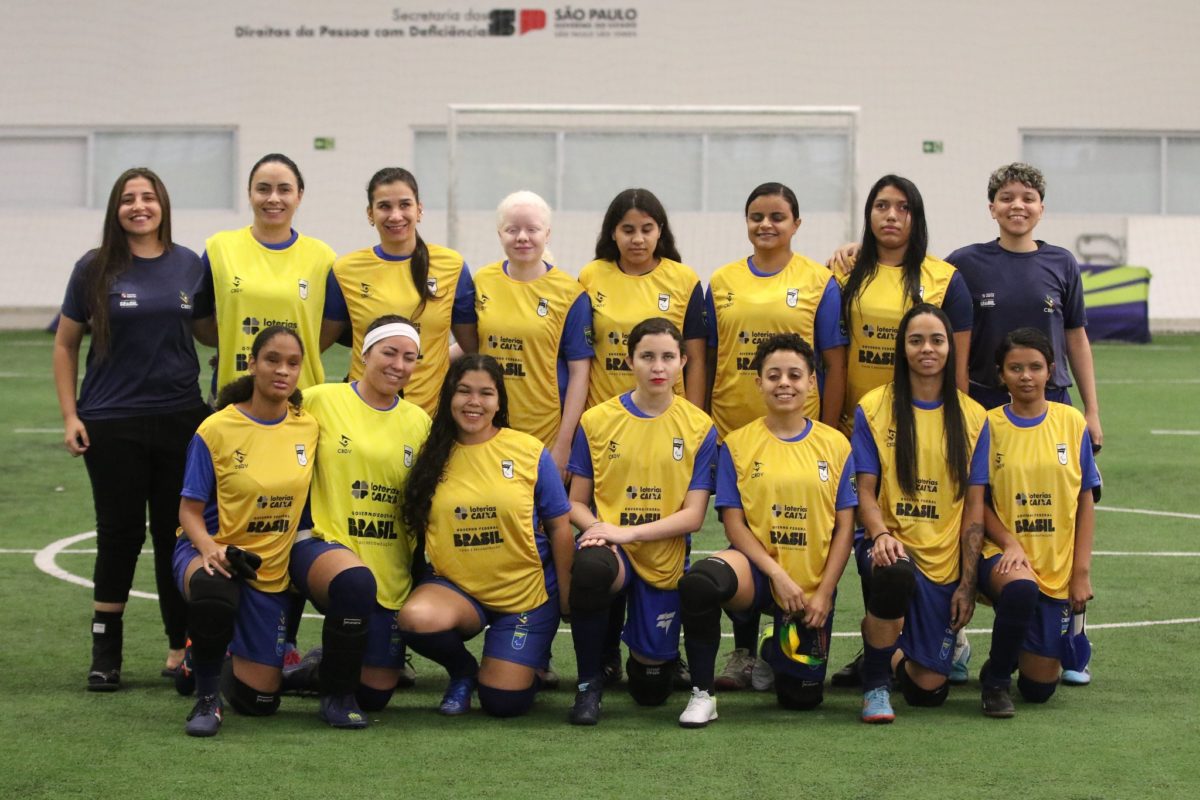 Foto colorida de um grupo de mulheres em um campo de futebol coberto. Elas estão posando organizadas em duas fileiras: a fileira de trás está em pé e a da frente está ajoelhada. A maioria das mulheres veste uniforme esportivo com colete amarelo, com o logo da Loterias Caixa, Governo Federal e Brasil estampados. Algumas usam faixa na cabeça e joelheiras. Nenhuma das atletas está com o óculos de proteção no momento da foto. No fundo, há um gol e uma parede branca com janelas e a inscrição "Secretaria dos Direitos da Pessoa com Deficiência - São Paulo". O ambiente é iluminado e o campo tem gramado sintético. Crédito: Renan Cacioli/ CBDV.