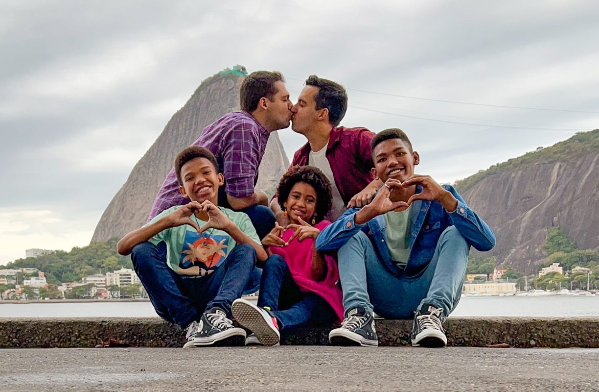 Foto colorida da família de Lucas e Carlos. Lucas e Carlos são dois homens brancos e estão se beijando, na frente deles, sentados, estão os três filhos, Kauã, Ketlin e Edgar, respectivamente. Eles são negros e fazem um desenho de coração com as mãos. Ao fundo, aparece o pão de açúcar