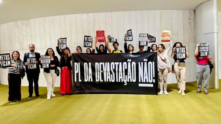 Foto colorida mostra pessoas em manifestação contra o PL da Devastação que implodiu o licenciamento ambiental no Brasil. Elas seguram uma faixa preta com letras brancas que diz: PL da Devastação não!