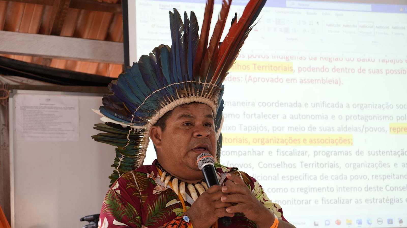 Foto colorida do cacique Gilson Tupinambá. Ele aparece com um cocar nas cores vermelho e azul e segura um microfone