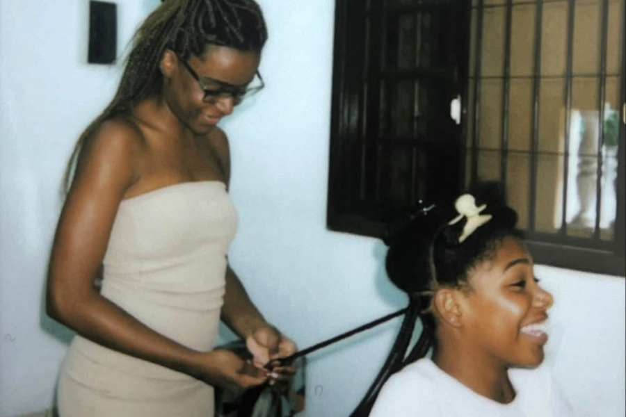 Foto colorida de Vitória fazendo tranças em outras mulher. Vitória começou a trançar para fazer seu próprio cabelo, já que não era acessível financeiramente fazer com profissionais. Faz sete anos que essa se tornou sua profissão (Foto: Arquivo pessoal)