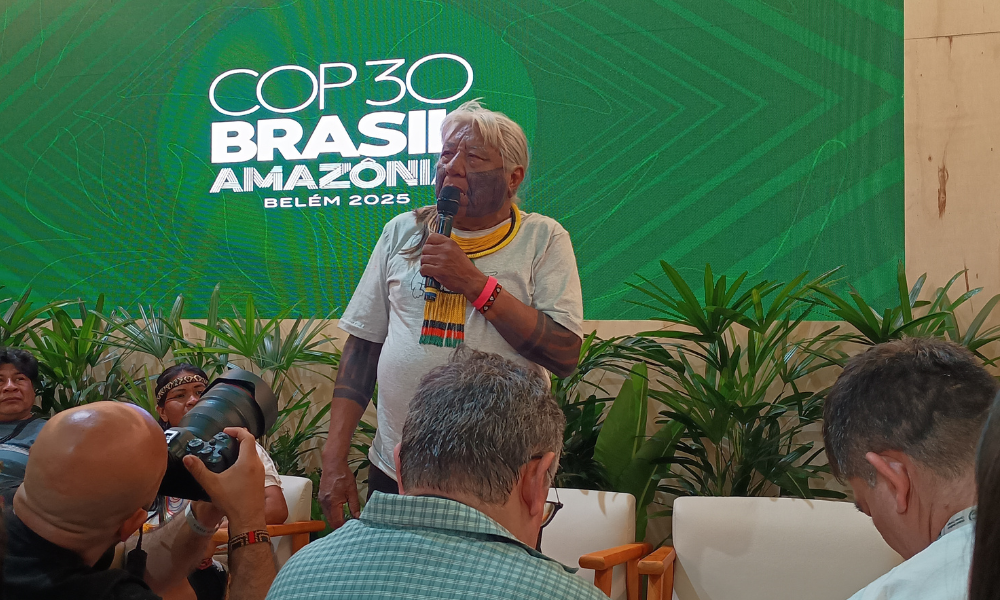 Foto colorida de Megaron Txucarramãe. Indígena de cabelos brancos e usa um colar do Instituto Raoni e segura um microfone. Ao fundo, painel da COP30