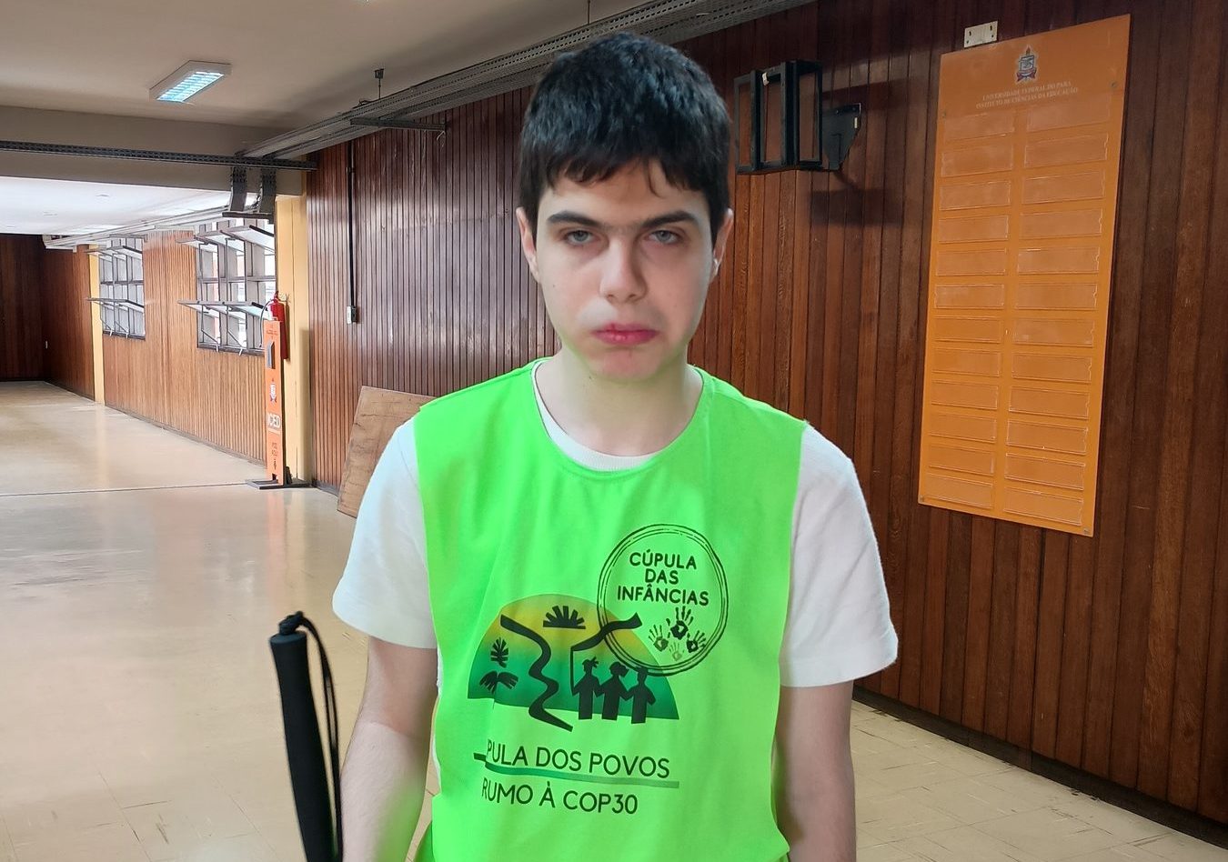 Foto colorida de Artur De Mari. Ele é um adolescentes branco, cego e com cabelo preto curto. Ele usa um colete verde da Cúpula dos Povos e segura uma bengala. Ao fundo, parede marrom