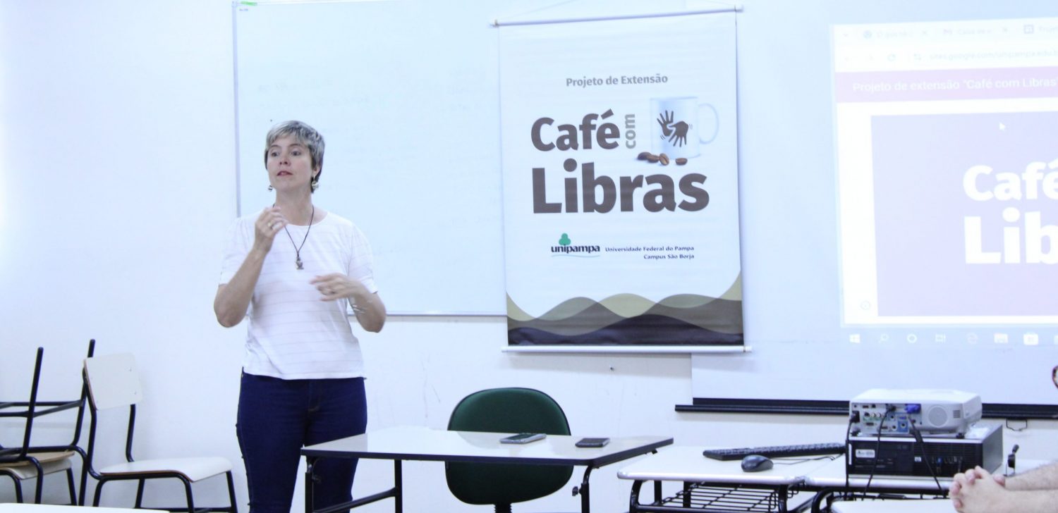 Foto colorida de Keli Krause em sala da Unipampa. Keli é uma mulher branca com cabelo curto e loiro. Ela usa uma camiseta branca e calça jeans. Ela gesticula. Ao fundo, cartaz do projeto Café com Libras