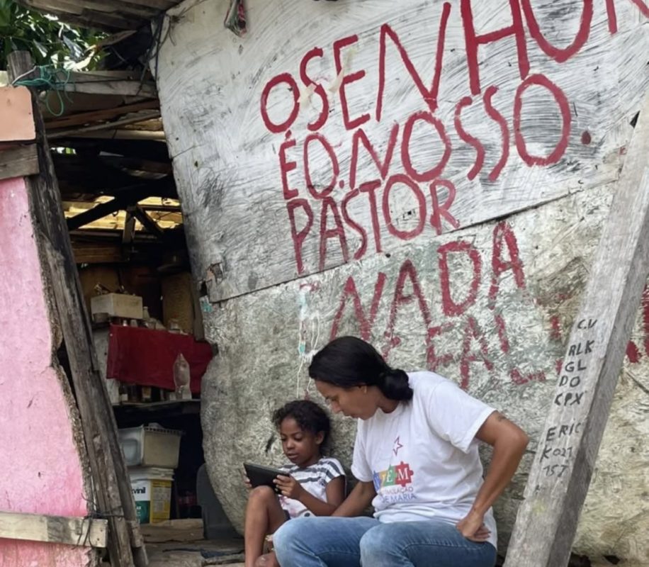 Foto colorida de Rafael França ao lado da filha. Ela é uma mulher negra com cabelo preto e está sentada, olhando para a filha, uma criança negra que segura um celular. Ao fundo, parede em que se lê: "o senhor é o nosso pastor nada falta"