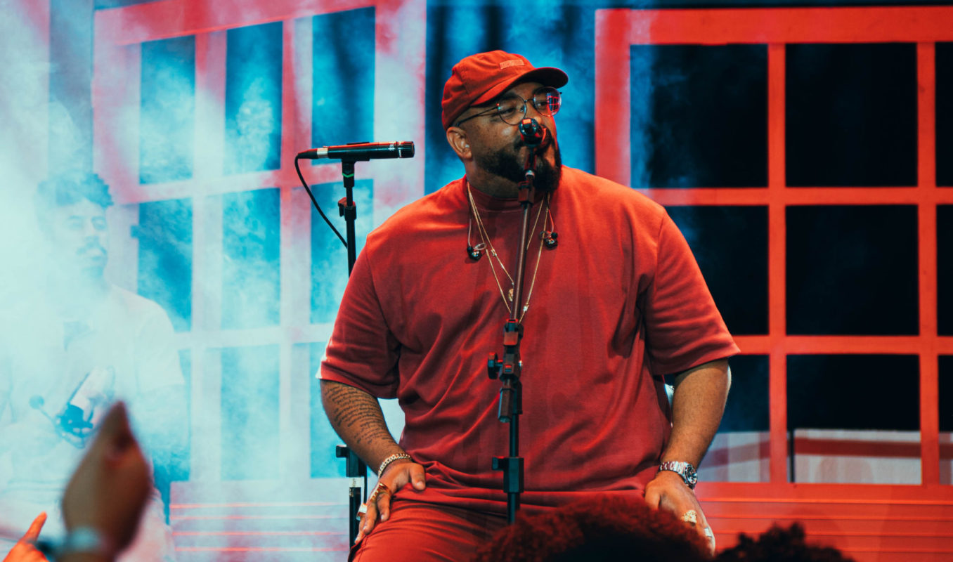 Foto colorida do cantor Tiee. Ele é um homem negro, com barba preta. Ele usa um bone e camiseta vermelha e está sentado em uma cadeira em um palco, com microfone diante dele