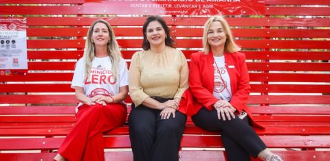 Foto colorida de três mulheres sentadas em um banco vermelho gigante em uma praça. Na imagem, as três aparecem olhando para a foto