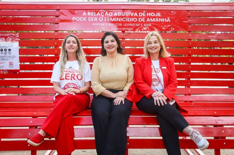 Foto colorida de três mulheres sentadas em um banco vermelho gigante em uma praça. Na imagem, as três aparecem olhando para a foto