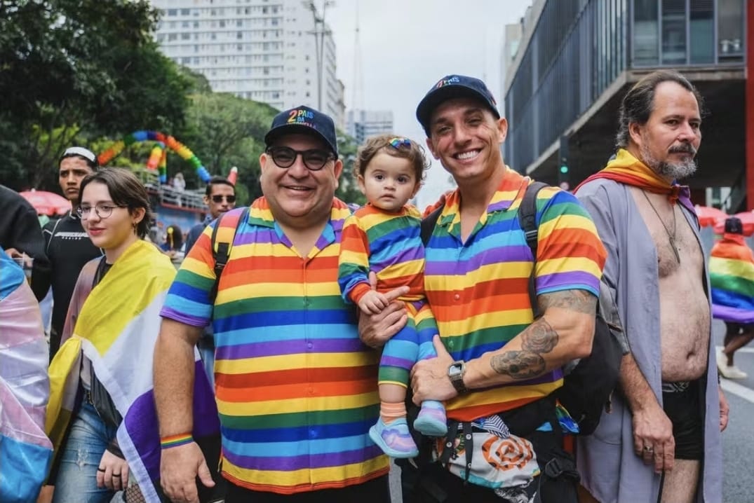 Foto colorida de Jarbas e Mikael com a filha Antonella no colo durante parada LGBT de São Paulo. Eles sorriem e usam roupas nas cores do arco-íris