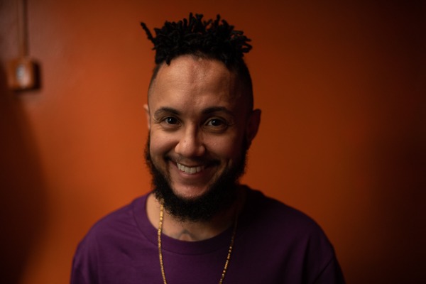 Foto colorida de Noah Scheffel. Ele é um homem egro com cabelo black e está sorrindo. Ele usa uma camiseta roxa e um colar amarelo. Ao fundo, parede laranja