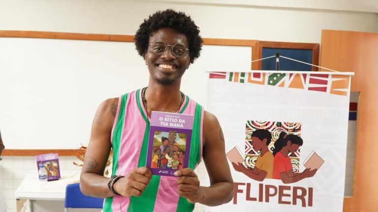 Foto colorida de Anderson Shon, homem preto com cabelo curto afro e segurando o livro Sitio da Tia Naná