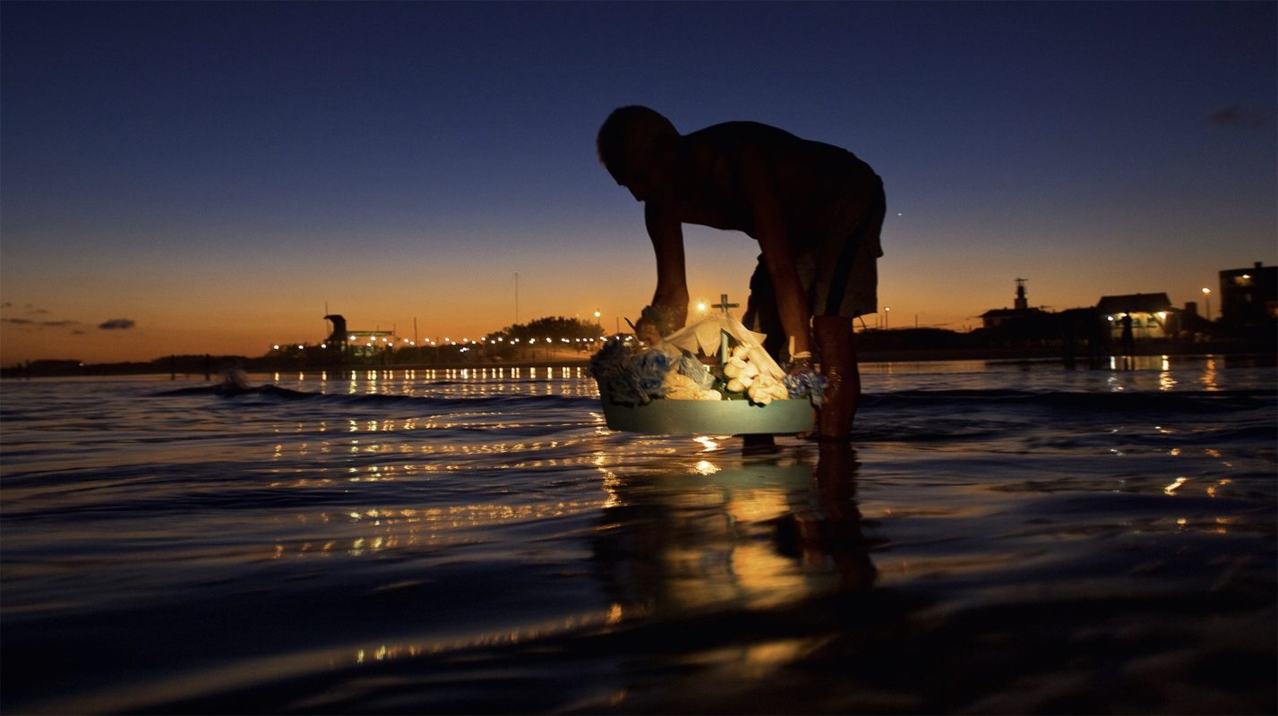 Foto colorida que mostra pessoa colocando barco de oferenda no mar. Batuque e tradições afro-gaúchas no Carnaval da Portela