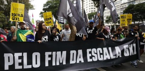 Foto colorida com manifestantes durante o grito dos excluídos no Rio de Janeiro. Eles seguram uma faixa preta com a frase, em branco: "Pelo fim da escala 6x1"