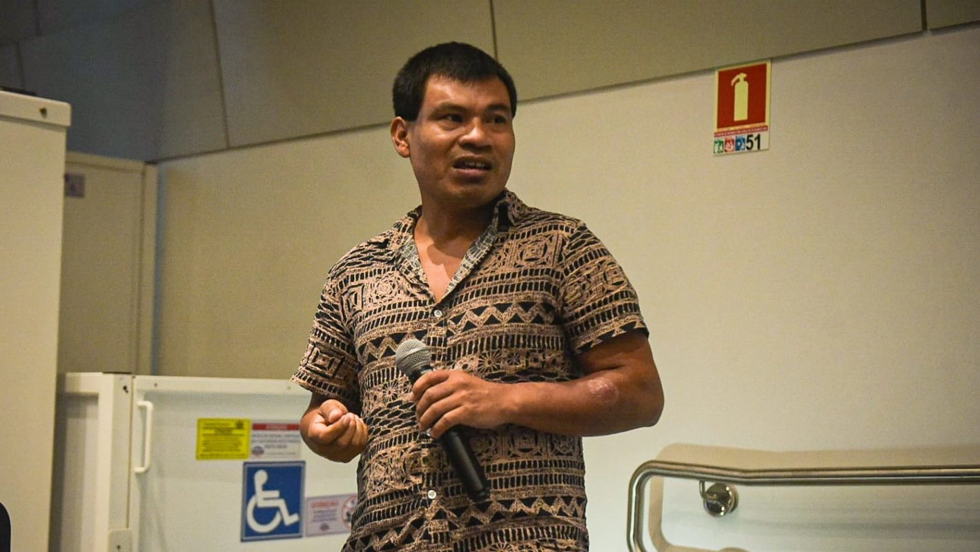 Foto colorida de Arnildo Werá Moreira durante reunião de lideranças indígenas com o MPF para tratar da fábrica de celulose. Ele é um indígena guarani com cabelo curto e segura um microfone