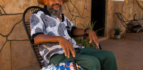 Foto colorida de Nego Bispo. Ele é um homem negro, com barba cheia e grisalha. Ele está sentado e usa uma camiseta branca com detalhes cinzas e um chapéu marrom