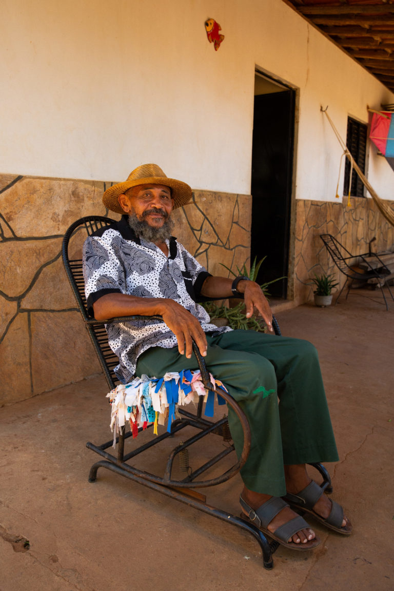 Foto colorida de Nego Bispo. Ele é um homem negro, com barba cheia e grisalha. Ele está sentado e usa uma camiseta branca com detalhes cinzas e um chapéu marrom