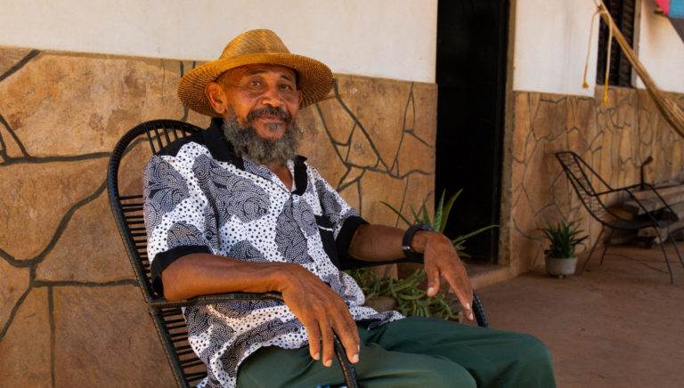 Foto colorida de Nego Bispo. Ele é um homem negro, com barba cheia e grisalha. Ele está sentado e usa uma camiseta branca com detalhes cinzas e um chapéu marrom