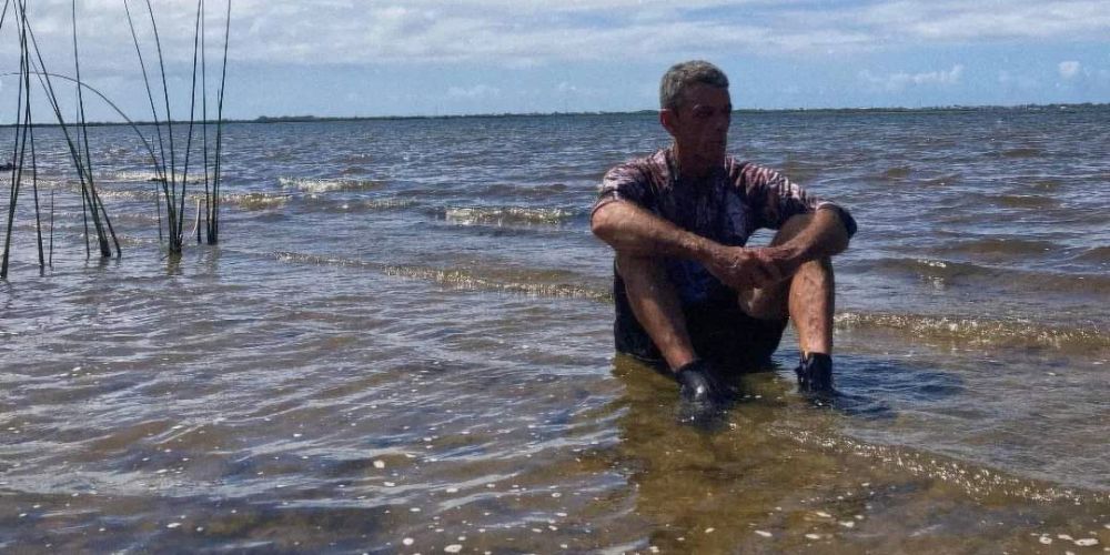 Foto colorida de Jorge Rosa sentado nas margens do Rio Tramandaí. Ele está com uma expressão série e olha para o lado