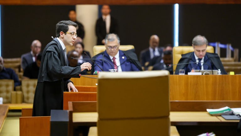 Foto colorida do Advogado da União João Pedro Carvalho, durante sessão do STF sobre a moratória da soja. Ele aparece no canto esquerdo da imagem, em pé, vestido com uma toga e próxima da tribuna com microfone