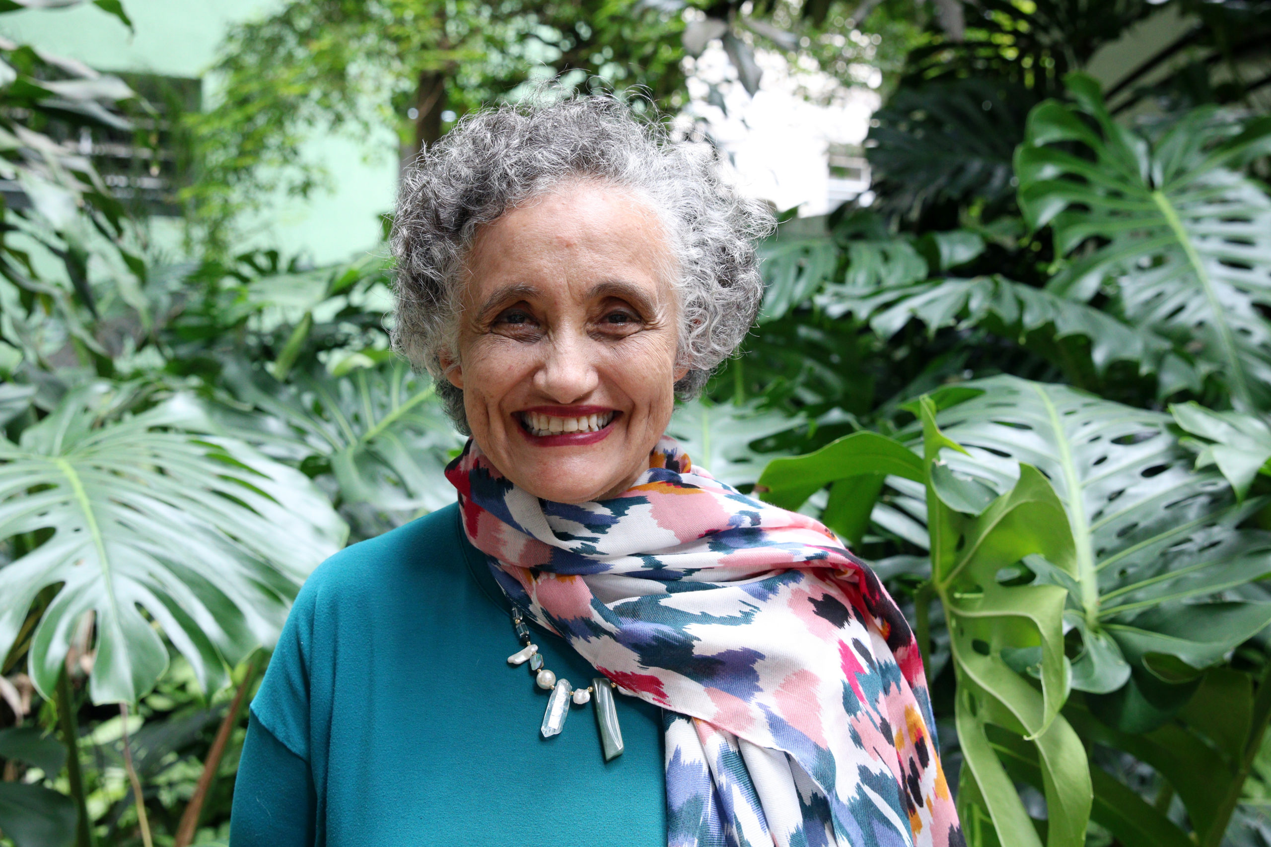 Imunologista Ester Sabino sorri em frente a plantas, em retrato ao ar livre.