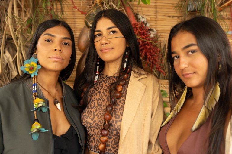 Foto colorida de Maiara Astarde, Vicka Tupi e Karol Kaysá. Criadoras do Sementes Urbanas, as três são mulheres indígenas e aparecem uma ao lado da outra, vestidas com adereços que remetem à sua cultura