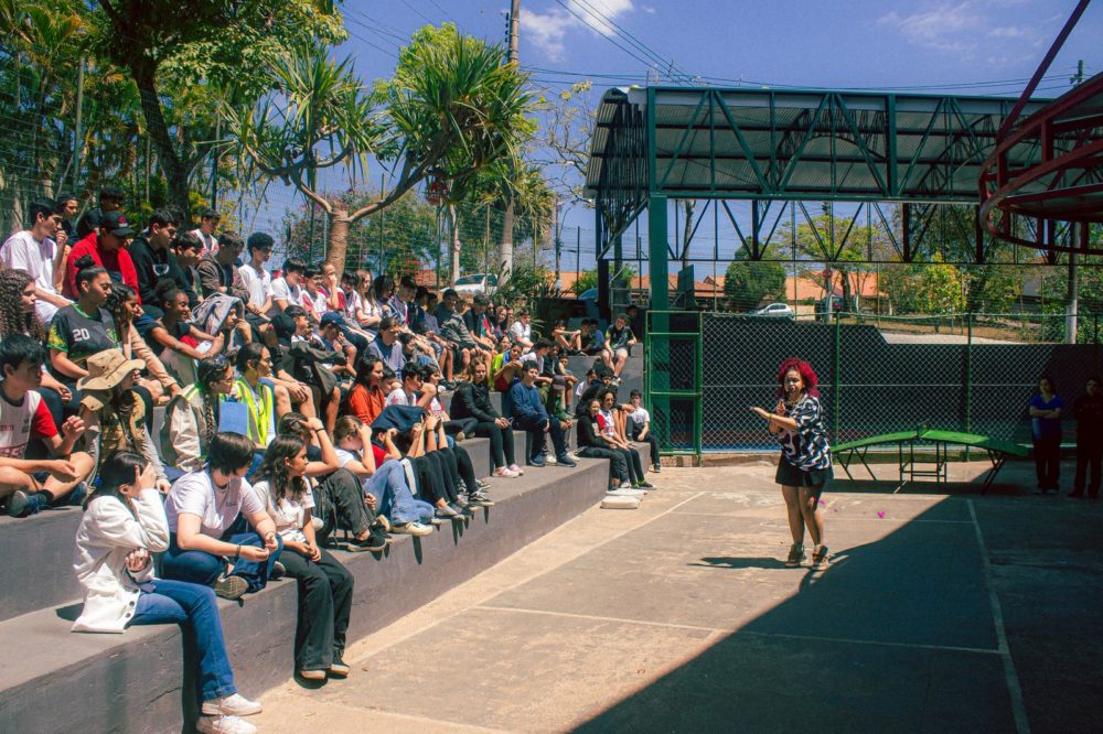 Foto colorida que mostra Mileny no canto direito em pé, diante de um grupo de jovens em uma arquibancada no canto esquerdo