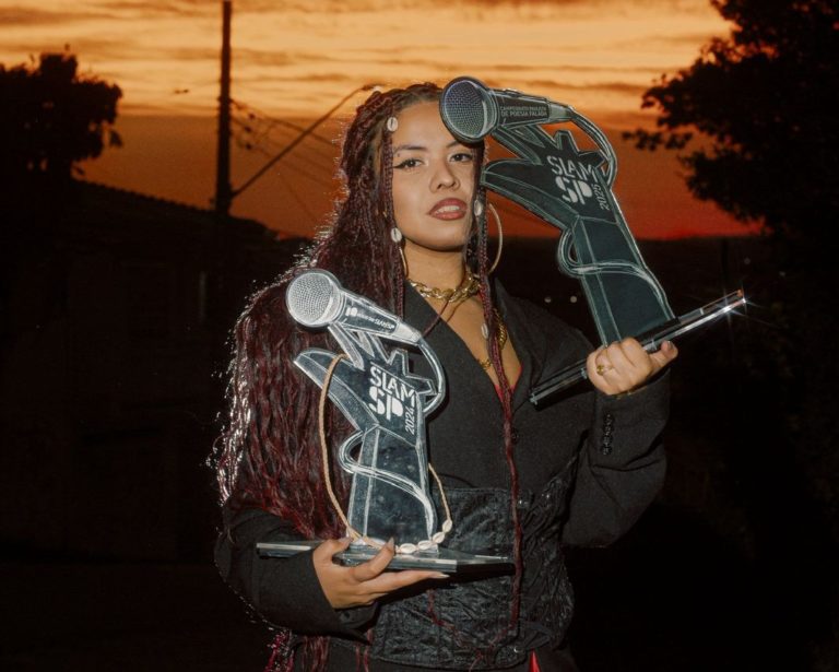 Foto colorida de Mileny Vitória. Ela é uma mulher negra, com cabelo afro em tranças e vermelho. Ela segura dois troféus e usa um casaco preto. Ao fundo, pôr do Sol