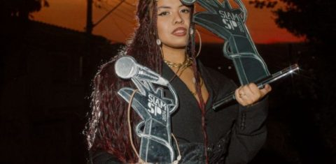 Foto colorida de Mileny Vitória. Ela é uma mulher negra, com cabelo afro em tranças e vermelho. Ela segura dois troféus e usa um casaco preto. Ao fundo, pôr do Sol