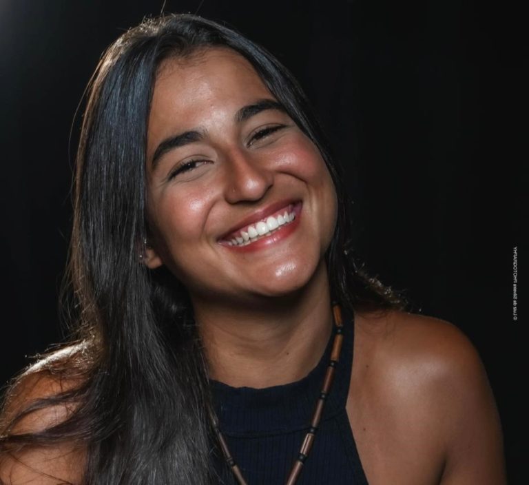 Foto colorida de Karol Kayá. Ela é uma mulher indígena, com cabelo preto liso. Ela está sorrindo e usa uma blusa preta com um colar. Fundo preto