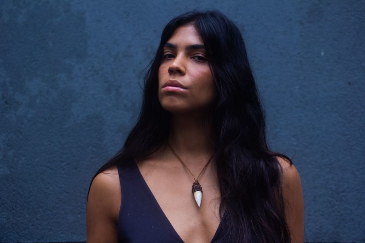 Foto colorida de Vicka Tupi. Ela é uma mulher indígena, de cabelo preto, longo, ela olha para a frente com expressão série. Ela usa uma blusa azul, com um colar com detalhes brancos e pretos. Fundo azul