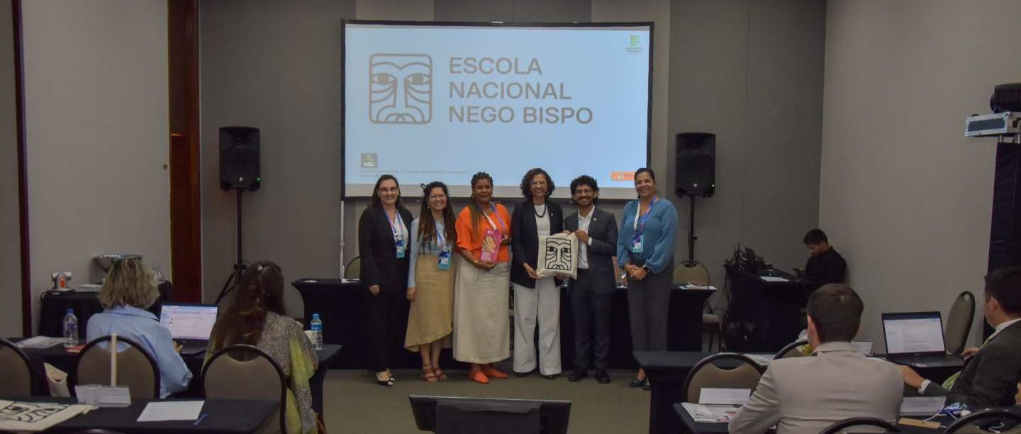 Foto colorida do lançamento do edital do Programa Nacional Escola Nego Bispo. Na imagem, um grupo de pessoas aparece em pé diante de uma tela com a lodo da iniciativa