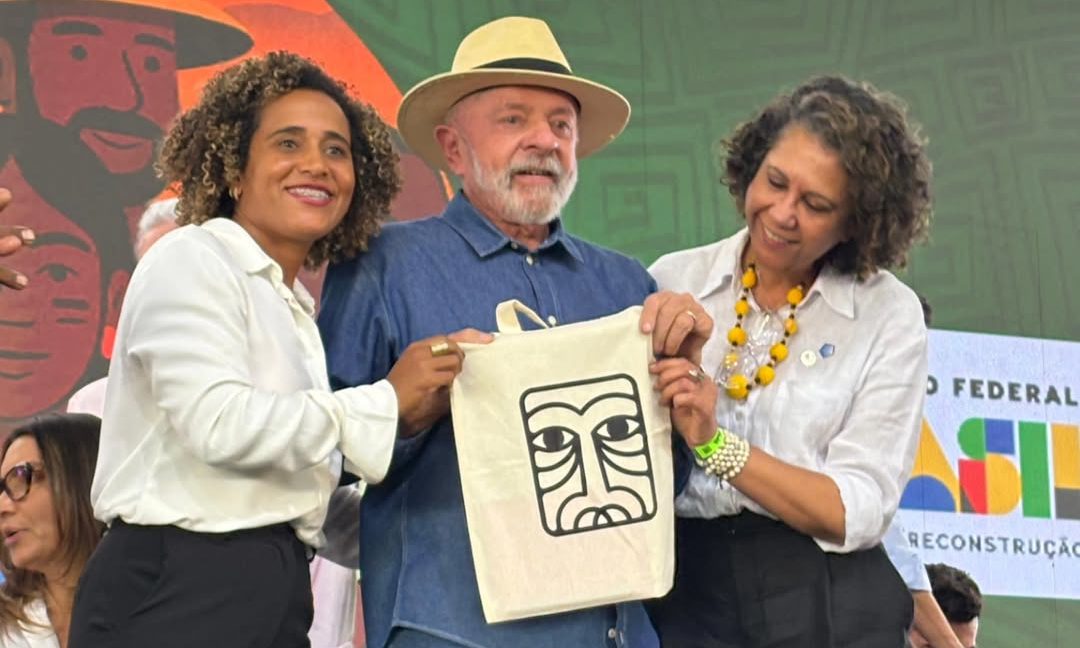 Foto colorida com Joana Maria, na esquerda, Lula, no centro, e Luzia Mota, na direita. Lula segura uma ecobag com o símbolo da Escola Nego Bispo.