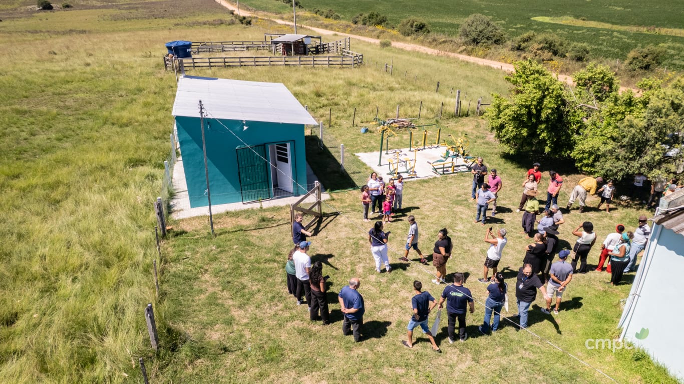 Foto colorida com vista aérea da agroindústria e de membros da comunidade de Ibicuí da Armada reunidos perto do prédio, pintado de azul com telhado de zinco
