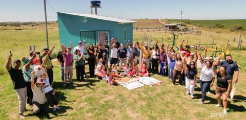 Foto colorida com várias pessoas reunidas em um semi-círculo no Quilombo Ibicuí da Armada, em Santana do Livramento. As pessoas estão com os braços erguidos e sorriem. Ao fundo, aparece a casa em que fica a agroindústria para apicultura e panificados