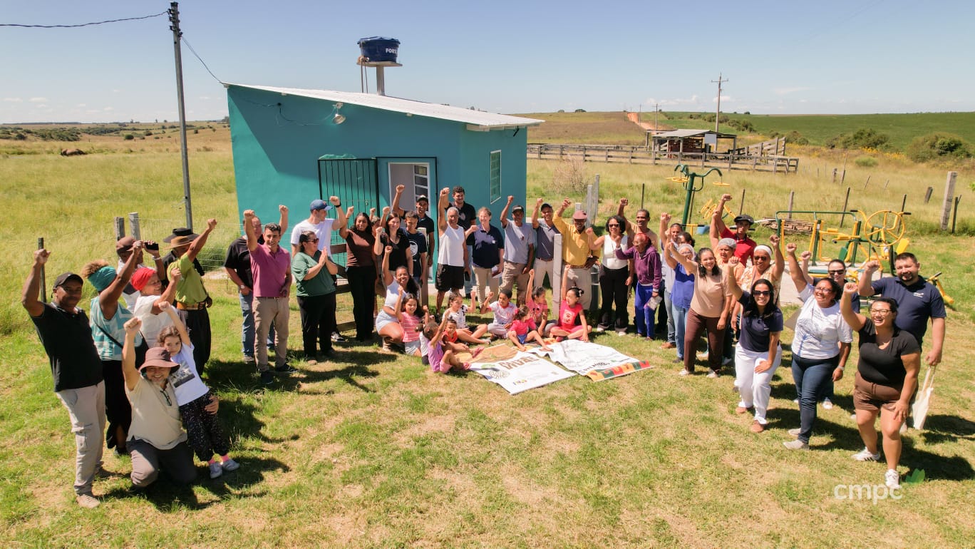 Foto colorida com várias pessoas reunidas em um semi-círculo no Quilombo Ibicuí da Armada, em Santana do Livramento. As pessoas estão com os braços erguidos e sorriem. Ao fundo, aparece a casa em que fica a agroindústria para apicultura e panificados