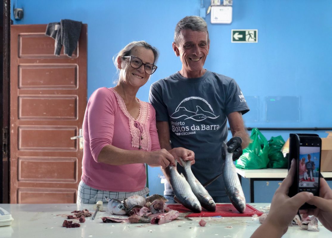 Foto colorida de Jorge Rosa ao lado da esposa. Ele é um homem branco, com cabelo branco e usa uma camiseta azul do Projeto Botos da Barra. Ela também é uma mulher branca e está usando uma blusa rosa. Ambos sorriem e seguram peixes diante de uma mesa